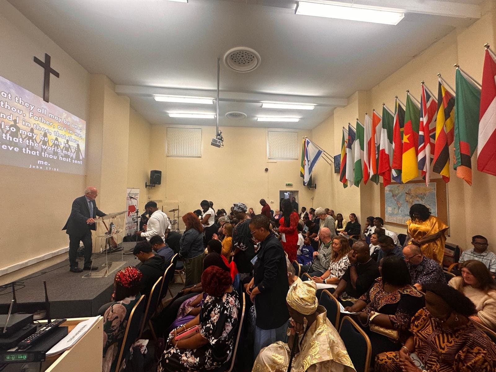 Pastor Malcolm preaching to a full congregation at King's Christian Centre, with flags of many nations along the wall